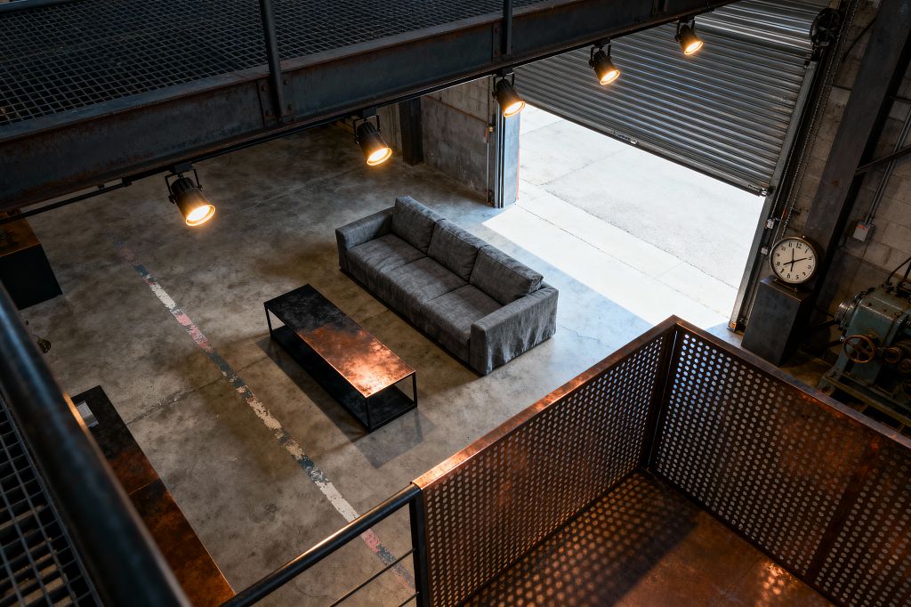 A double-height industrial living room viewed from an upper steel mezzanine shows a gray three-seat sofa paired with a long, low metal coffee table on a sealed concrete floor marked by a faded painted stripe. Warm spotlights hang from the catwalk while daylight pours through a partially open corrugated roll-up door. Exposed concrete block walls, a perforated steel railing with rust patina, visible conduit, and a large analog clock emphasize the raw warehouse character.
