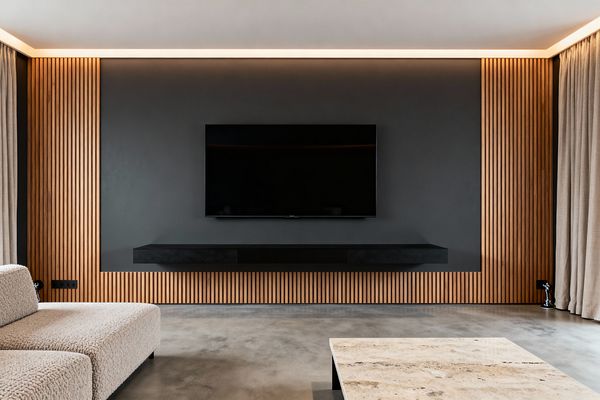 A minimalist media room features a charcoal-gray media wall bordered by vertical slatted oak, holding a wall-mounted flat-screen TV above a slim matte-black floating console. The space includes a polished concrete floor, a low cream bouclé sectional, a beige travertine coffee table with dark legs, full-height linen curtains on the right, warm perimeter cove lighting at the ceiling, and small black floor spotlights for subtle accents.
