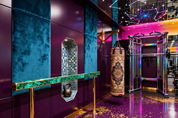 A jewel-toned home gym features glossy plum walls with large teal velvet panels and an arched blue mosaic tile niche displaying a kettlebell. A malachite-pattern barre on polished brass posts faces a textile-wrapped punching bag and an iridescent chrome cable machine. Overhead, a mirrored ceiling with constellation-style LEDs and magenta-to-amber lighting reflects a purple epoxy floor sprinkled with gold flecks.