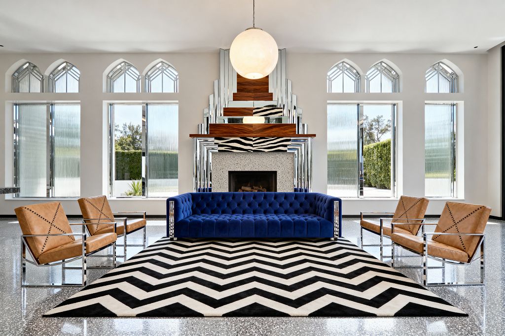 A symmetrical Art Deco living room shows a deep blue tufted velvet sofa on a black-and-white chevron rug, centered before a stepped mirrored fireplace with a terrazzo surround, walnut mantel, and a zebra-pattern accent. Four camel leather armchairs with chrome frames face the sofa on a speckled terrazzo floor. Tall arched windows with ribbed glass line white walls, and a large opaline globe pendant hangs above, brightening the space.