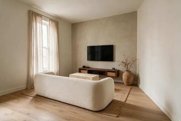 A serene media room with a wall-mounted black TV centered on a textured beige accent wall, a low wooden media console with woven storage baskets beneath it, and a cream boucle sofa facing the screen. A jute rug, pale wood floors, sheer linen curtains, and a large ceramic vase with dried branches add warm, natural texture.