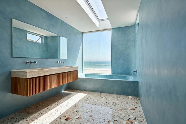 A bright coastal bathroom with sea-glass blue polished plaster walls and a built-in tub in the same finish. A floating ribbed teak vanity with a light travertine countertop holds two integrated sinks and wall-mounted metal faucets beneath a wide mirror. The terrazzo floor is inlaid with shell and stone chips. Daylight streams through a skylight and a tall fluted window that frames an ocean view, creating a calm, airy atmosphere.