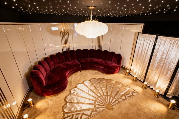 A glamorous media room features a curved, scalloped-back sectional sofa in deep burgundy velvet on a pale, plush carpet with a fan motif in satin inlay. Glossy ivory wall panels with gold Art Deco lines wrap the room. A large white frosted-glass pendant hangs beneath a star-like ceiling, while brass floor uplights illuminate the walls. A white damask folding screen stands to the side before black drapery.