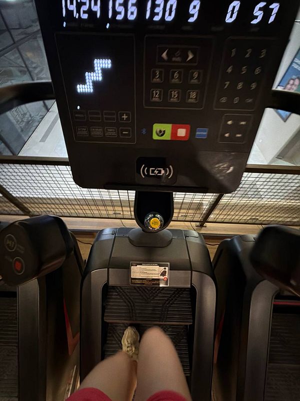 A woman in gym attire is exercising on a treadmill inside a gym with large windows overlooking a cityscape.