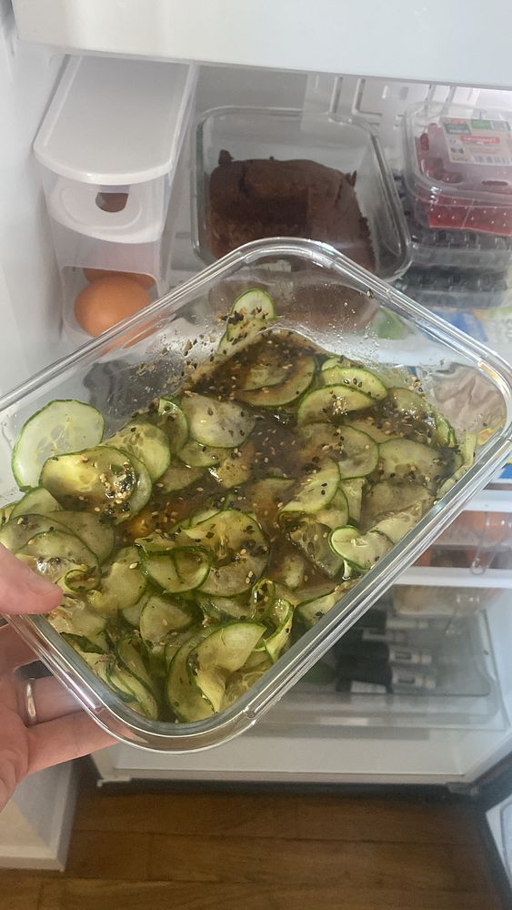 A container of sliced cucumbers in a marinade is being held in front of an open refrigerator.