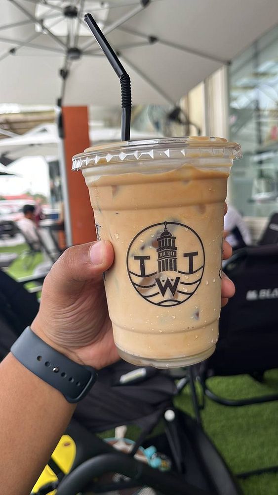 A hand holds a cup of iced coffee with a logo, set in an outdoor café environment.
