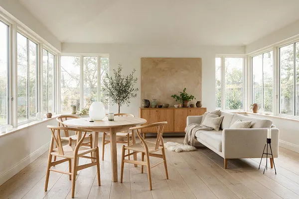 A bright Scandinavian-style sunroom with wraparound windows, pale wood floors, a round oak dining table, four matching wooden chairs, and a light neutral sofa. A wooden sideboard with ceramics and plants sits against the back wall, and soft beige textiles add warmth to the minimalist space.