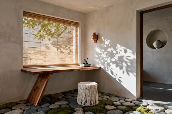 A serene wabi-sabi home office features a floating live-edge wood desk fixed to pale lime-plaster walls beneath a large window with a bamboo shade, casting dappled leaf shadows across the room. The floor is made of rounded river stones with patches of moss and a white, fringed rope stool; a terracotta wall sconce, a small stone tray with pencils on the desk, and a doorway revealing a recessed niche with a weathered ceramic jar complete the natural, minimalist setting.