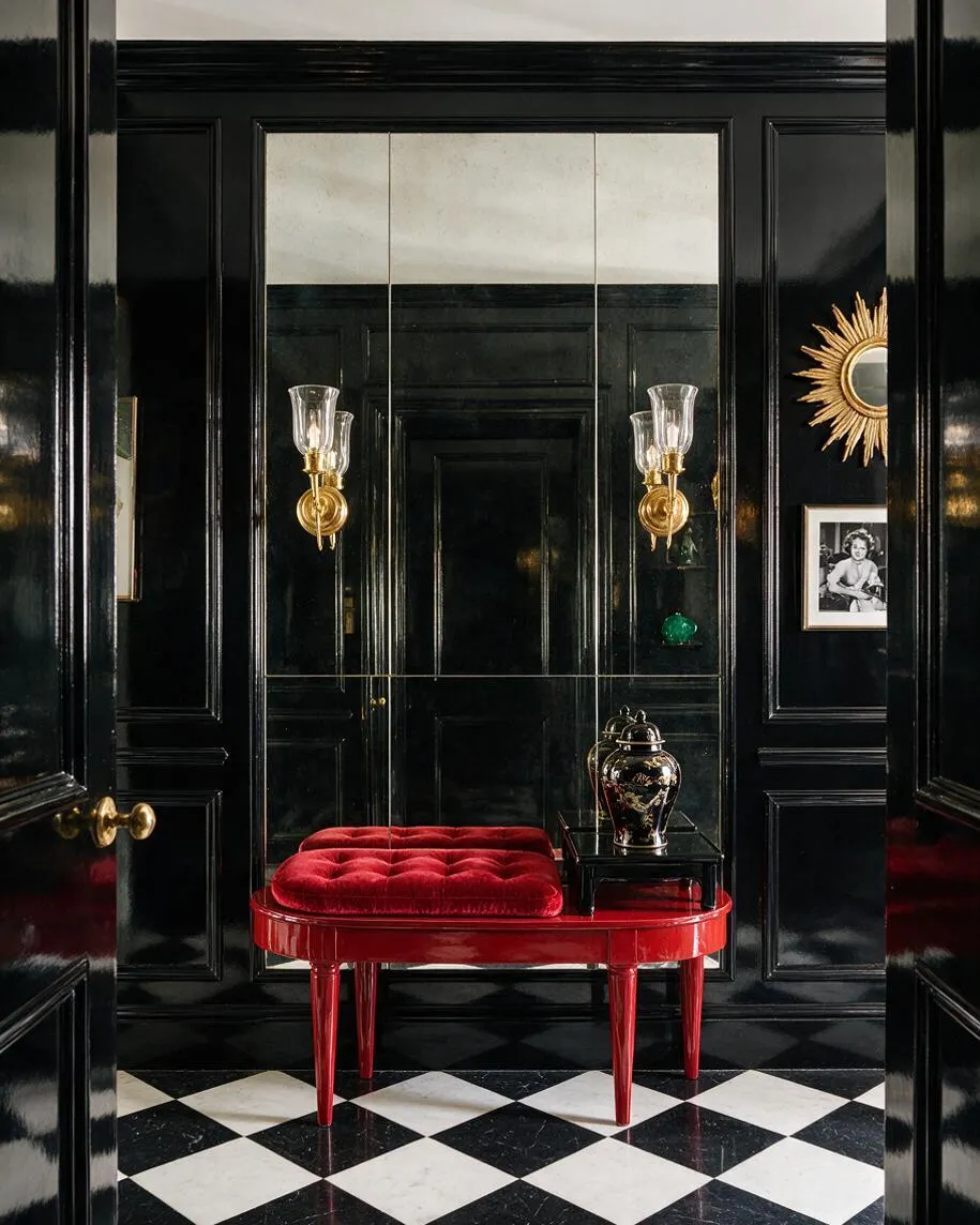 A dramatic Hollywood Regency hallway with glossy black paneled walls, a black-and-white checkered floor, and a bright red lacquer bench topped with a tufted velvet cushion. Gold sunburst accents, brass wall sconces, a mirrored back wall, and a small black side table with a decorative urn add glamour and contrast.