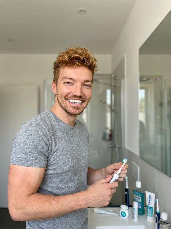 A smiling man stands in a modern bathroom holding a toothbrush.