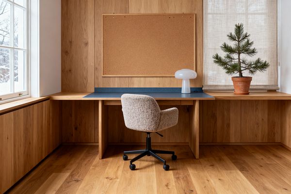 A Scandinavian home office with warm oak wall paneling and matching plank floors centers on a built-in corner desk, its work area defined by a muted blue laminate top and slim backsplash. A large cork pinboard hangs above, a glossy white mushroom-shaped lamp sits to the right, and a small pine tree in a terracotta pot rests before a linen roller shade; a beige bouclé swivel chair with a black wheeled base faces the desk, lit by natural light from a mullioned window on the left.