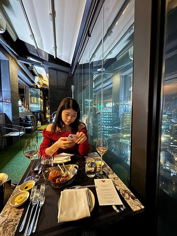 A woman is seated at a beautifully set table on a rooftop restaurant, engrossed in her phone with a stunning city view behind her.