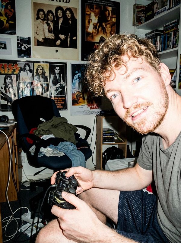 A man with curly hair and a beard is sitting in a cluttered room, holding a video game controller.