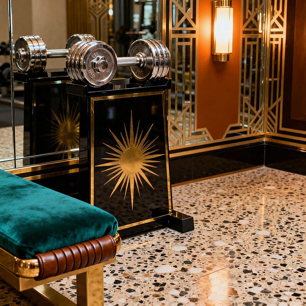 An Art Deco home gym corner features chrome dumbbells resting on a black lacquer pedestal adorned with a gold sunburst, reflected in mirrored walls with geometric brass detailing. In front, an emerald velvet bench with a brass base and brown leather-wrapped end sits on speckled terrazzo flooring, while a brass sconce with a frosted glass shade warms terracotta wall panels and glossy black trim.