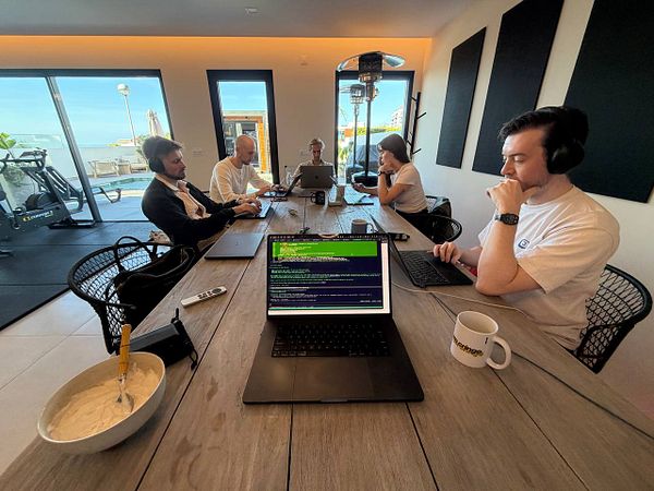 Five people working together at a long table in a modern indoor coworking space with laptops and headphones.