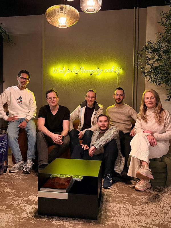 A group of six people poses together in a cozy indoor setting with a neon sign in the background.