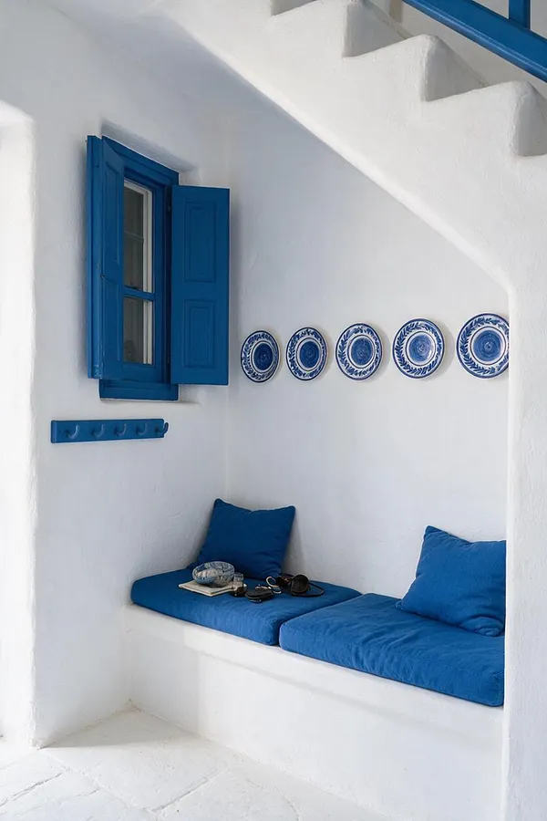 A compact Mediterranean-style mudroom nook with white plaster walls, a bright blue shuttered window, and a built-in bench with vivid blue cushions and pillows. Decorative blue-and-white plates line the wall above the bench, and a matching blue coat hook rail adds function and color.