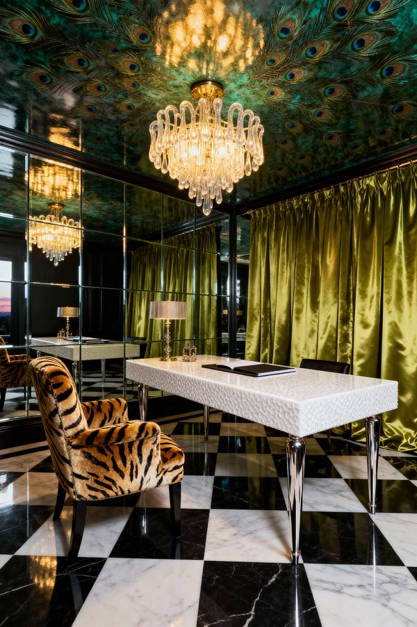 A glamorous Hollywood Regency home office shows a white textured desk on chrome legs, a plush tiger-print armchair, and a black-and-white marble checkerboard floor. One wall is lined with mirrored panels within black frames. Chartreuse satin curtains pool to the floor, and a glossy ceiling painted with peacock feathers reflects a crystal chandelier with gold detailing. A small crystal table lamp and minimal accessories sit on the desk, emphasizing light and reflection.