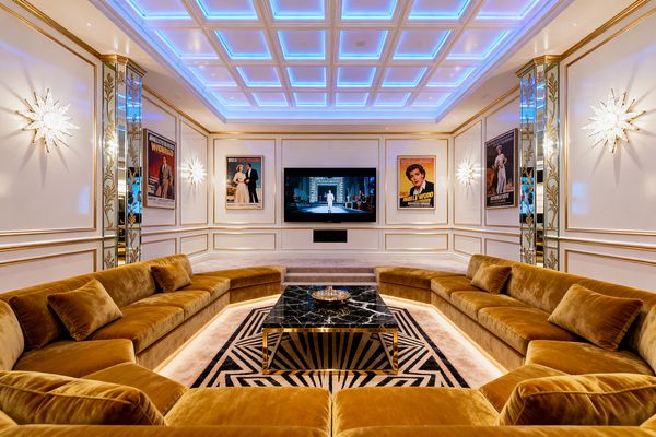 A glamorous media room features a U-shaped mustard velvet sectional surrounding a black marble coffee table with a brass base atop a black-and-white geometric rug. White paneled walls with gold trim are accented by mirrored pilasters, starburst crystal sconces, and framed classic movie posters, while a large wall-mounted TV centers the space. A coffered ceiling glows with blue LED lighting, and under-sofa illumination and plush carpet create a warm, cinematic ambiance.