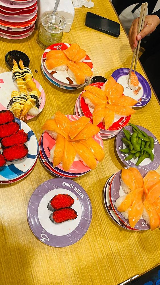 A close-up image of a person holding a piece of salmon sushi with chopsticks over a wooden table with a sushi platter and other food items.