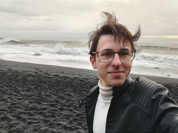 A person stands on a black sand beach with waves crashing in the background.