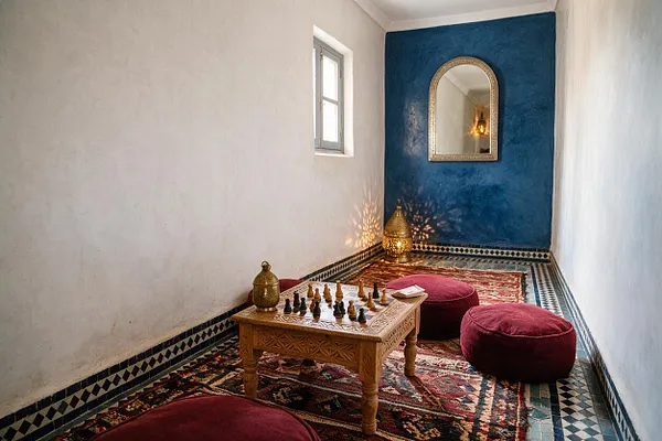 A narrow Moroccan-style game room with white plaster walls, a deep indigo accent wall, and patterned mosaic trim along the floor. The room features layered red rugs, burgundy floor cushions, a carved wooden chess table with pieces arranged on top, a brass lantern, and an arched mirror that reflects warm light.