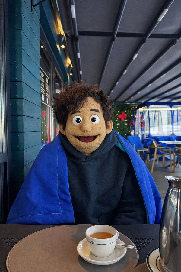 A puppet character is seated at a restaurant table, wearing a blue blanket and a black hoodie, with a cup of tea in front of it.