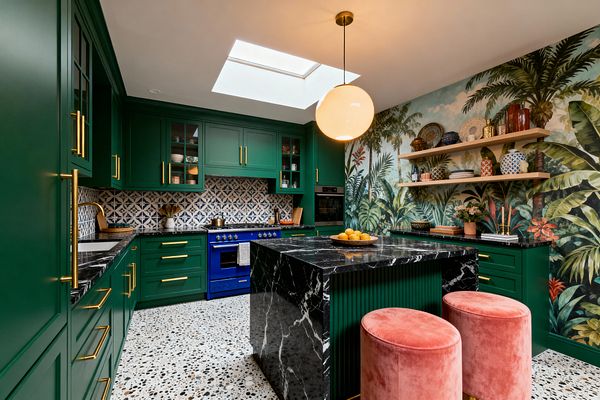 A maximalist kitchen features emerald green Shaker-style cabinets with brass hardware, black marble countertops, and a cobalt blue range set beneath a blue-and-white geometric tile backsplash. A skylight and globe pendant illuminate a terrazzo floor, a fluted black-marble waterfall island topped with lemons, blush pink velvet stools, and a full-wall tropical palm mural with two light wood shelves displaying colorful ceramics and plants.