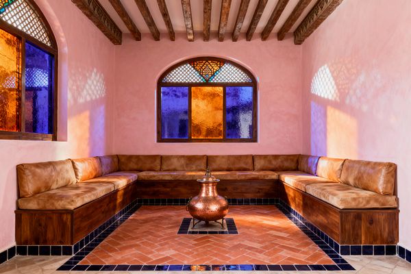 A Moroccan-style living room with blush pink plaster walls and exposed wooden ceiling beams features a U-shaped built-in wooden banquette topped with camel-colored leather cushions. The floor is terracotta laid in a herringbone pattern with a black tile border, and a hammered copper urn sits at the center. Arched windows with cobalt blue and amber stained glass and lattice details cast colorful light and patterned shadows across the room.