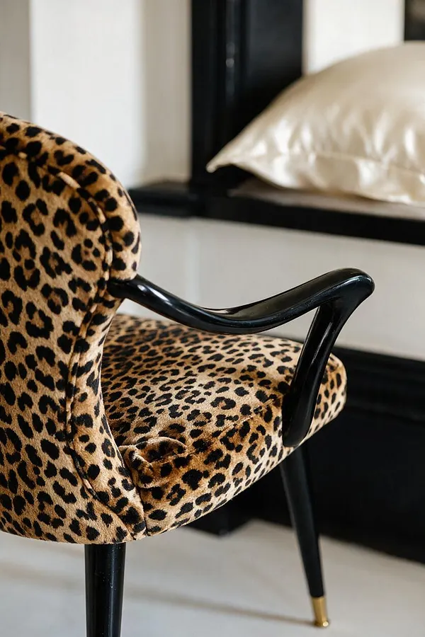 A close-up of a Hollywood Regency accent chair upholstered in leopard-print velvet with a rounded back, cushioned seat, and glossy black curved arms. The chair has slender black legs tipped with brass, set against a black-and-white interior with a cream satin pillow in the background.