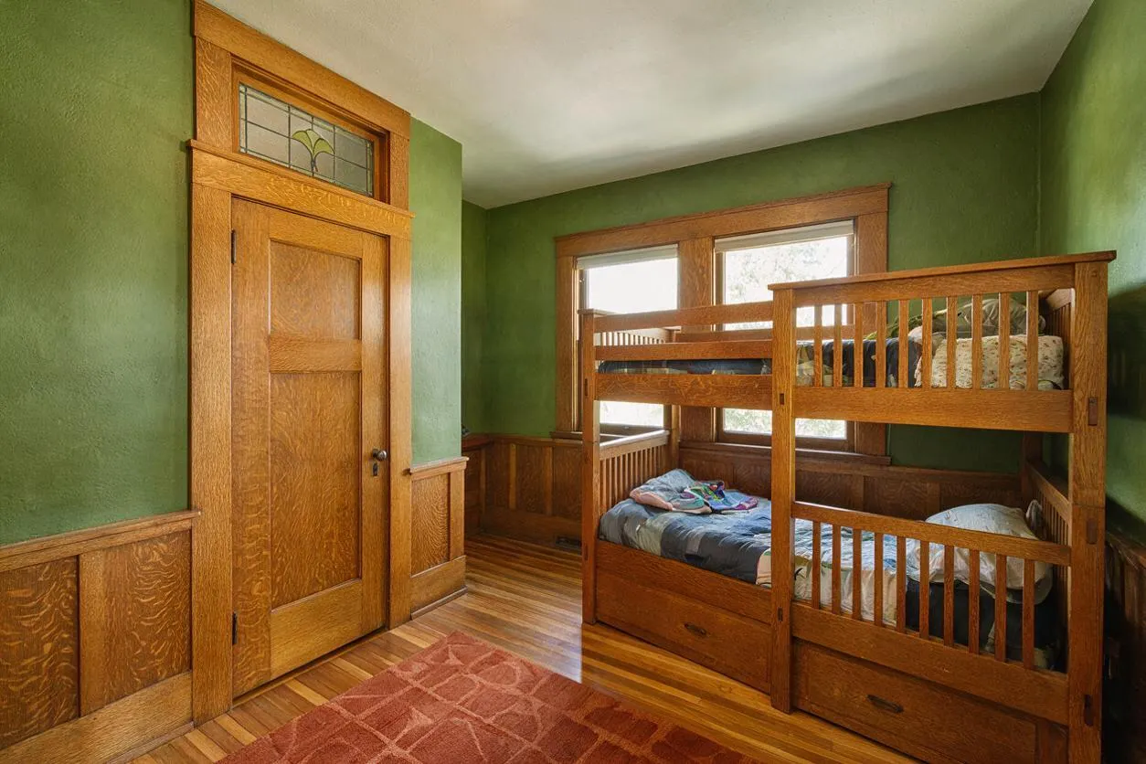 A Craftsman-style kids' room with green walls, warm wood wainscoting, and a built-in wooden bunk bed along the right side. A paneled wood door with a small stained-glass transom sits on the left, while windows above the bunk bed bring in natural light.