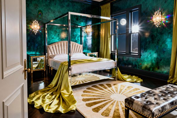 A glamorous bedroom shows a polished chrome four-poster bed with a scalloped cream velvet headboard, draped in chartreuse satin that spills onto an ivory rug with a gold sunburst. Emerald and teal marbled walls, crystal sconces, mirrored and gilded nightstands, black-trimmed windows with matching chartreuse curtains, a dark herringbone wood floor, and a tufted charcoal bench with nailhead trim complete the Hollywood Regency look.