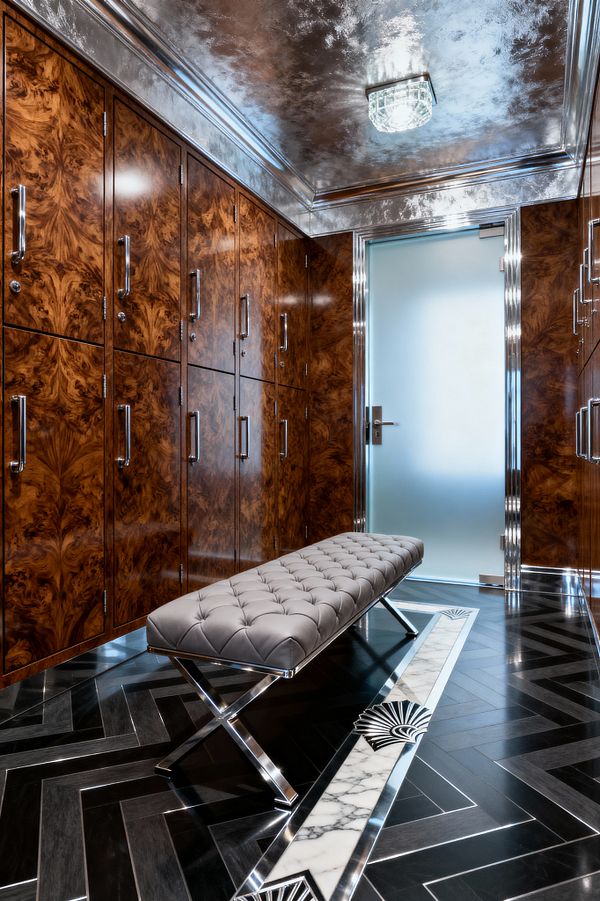 A luxurious mudroom lined with high-gloss burlwood lockers fitted with polished chrome pulls. A tufted gray leather bench with X-shaped chrome legs sits on a dark herringbone floor with a white marble inlay featuring black Art Deco fan motifs. The ceiling is finished in reflective silver leaf with a crystal flush-mount light, and a frosted glass door framed in chrome closes the space.