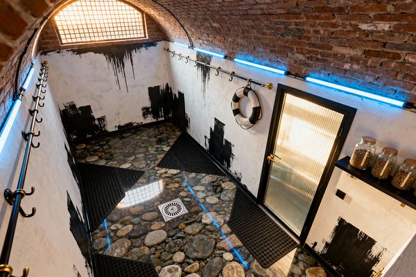 A small mudroom with a barrel-vaulted red brick ceiling and distressed white walls marked by black paint strokes. Steel rails with brass hooks line both sides, illuminated by blue tube lights. The floor is a glossy epoxy surface encapsulating rounded river stones with a central drain, topped by black rubber mats. A ribbed glass door with a black frame and brass handle faces a wall-mounted life ring and a shelf holding glass jars with cork lids.