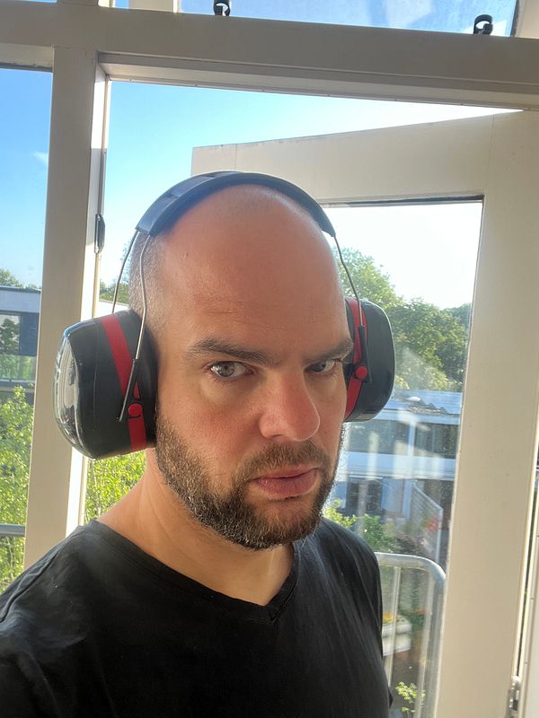 A man wearing protective headphones stands in front of a window with a view of greenery.