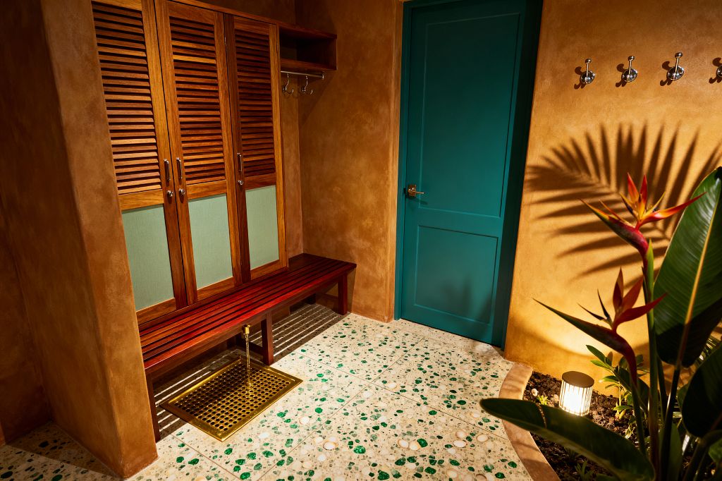 A tropical mudroom with textured ochre plaster walls features a teal door, a terrazzo floor with green and white chips, and a slatted teak bench in front of louvered lockers with pale green panels. A brass floor rinse tray sits beneath the bench, chrome hooks line the entry wall, and a bird-of-paradise beside a small uplight casts leafy shadows across the space.