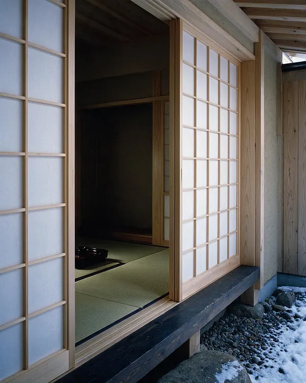 A Japanese Zen exterior showing a wooden facade with sliding shoji screens and an open entry revealing a dim tatami room inside. The structure sits above a dark base beam, with gravel, rocks, and patches of snow below, creating a calm, natural setting.
