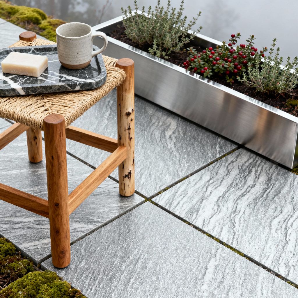 A Scandinavian patio vignette features large gray granite pavers with linear veining and moss between joints. In the foreground is a natural wood stool with a woven seat, topped by a black marble tray holding a speckled gray stoneware mug and a soap bar; along the edge, a brushed stainless-steel trough planter grows thyme and a small shrub with red berries against a soft, misty backdrop.