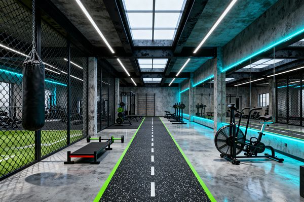 A long industrial-style gym with raw concrete walls and ceiling, polished concrete floors, and large skylights is lit by linear LED strips. A speckled black sprint track with neon green edges runs down the center, flanked by chain-link partitions and a full mirrored wall with cyan lighting. Black steel racks, benches, an air bike, and a hanging black heavy bag create a dark, high-contrast training zone.