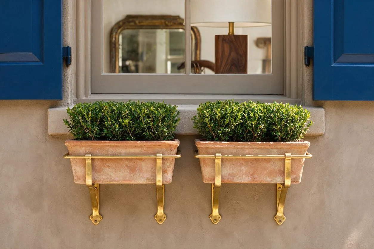 A close-up of a stucco exterior wall with a cream-framed window and bright blue shutters on either side. Two terracotta window boxes with brass brackets hold dense green shrubs beneath the sill, creating a colorful eclectic facade detail.