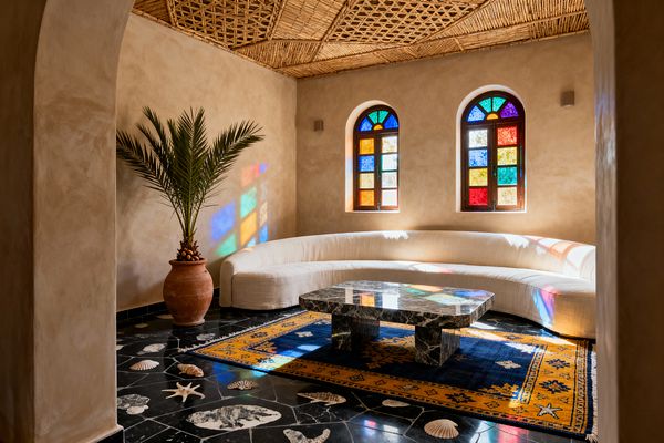 A sunlit Moroccan-style living room with smooth sand-colored plaster walls and a woven reed lattice ceiling. Two arched, wood-framed windows fitted with multicolored stained glass cast jewel-toned reflections onto a long curved ivory sofa. At center sits a chunky black marble coffee table on a deep blue rug with a saffron border, while a terracotta pot with a palm and scattered seashell decor rest on glossy black marble flooring.