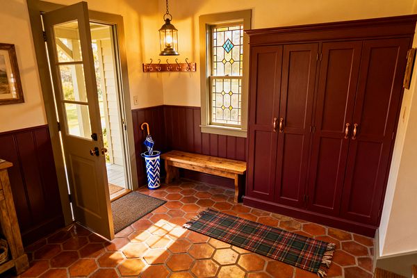 A traditional mudroom features terracotta hexagon floor tiles and a narrow tartan runner, illuminated by warm sunlight through an open sage-green door. Oxblood-painted wainscoting and matching Shaker cabinets with copper pulls line the wall beneath a leaded stained-glass window; a rustic wooden bench, a wood coat-hook rail, an antique-style lantern pendant, and a blue-and-white umbrella stand complete the space.