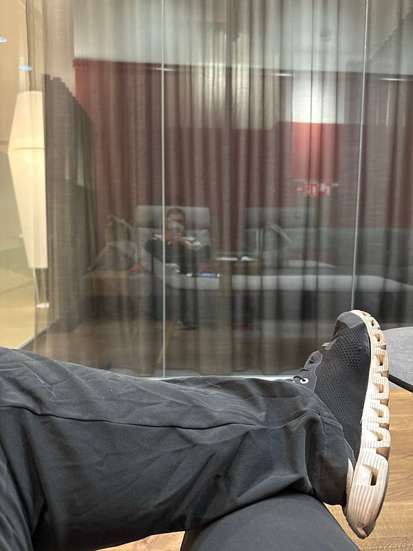 A person is relaxing in an airport lounge, with their legs crossed and a reflection visible in the glass partition.