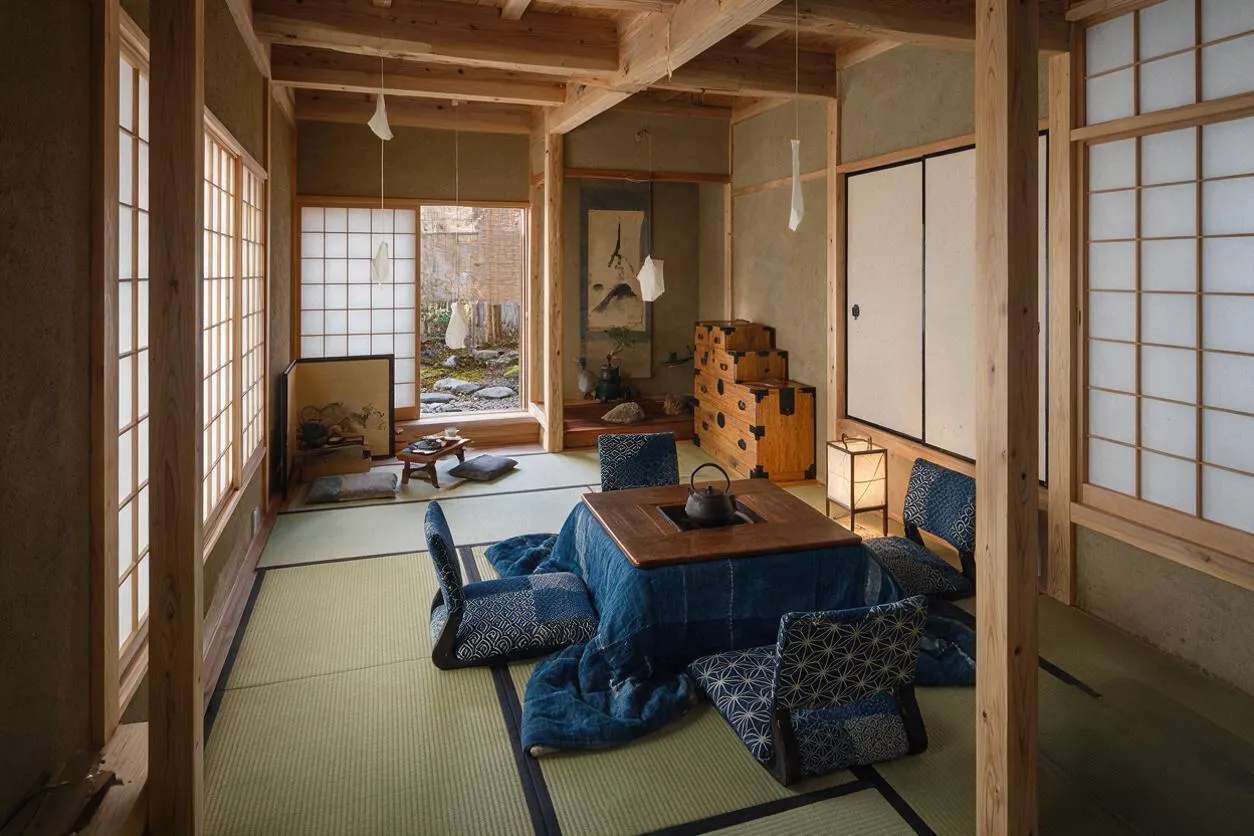 A tranquil Japanese Zen-style studio apartment with tatami mat flooring, exposed wooden beams, and shoji paper screens. Low blue floor cushions surround a small wooden table with a kettle, while a lantern, stacked chests, and simple wall art complete the calm, natural interior.