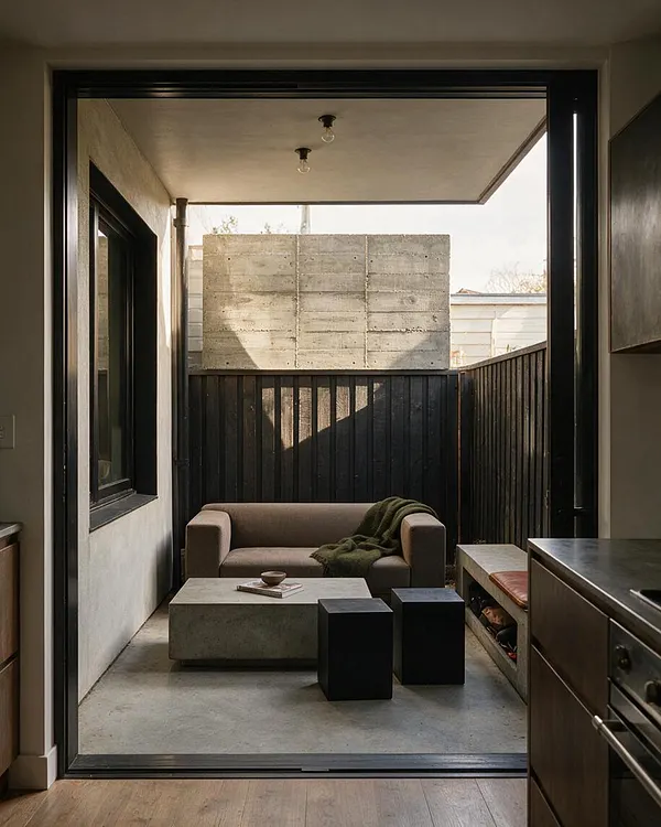 A narrow backyard lounge viewed through a large open doorway, featuring a raw concrete wall, dark vertical timber fencing, and a simple low sofa with a draped throw. A concrete coffee table and black cube stools sit on the concrete floor under a covered overhang, creating a minimal Brutalist outdoor living space.