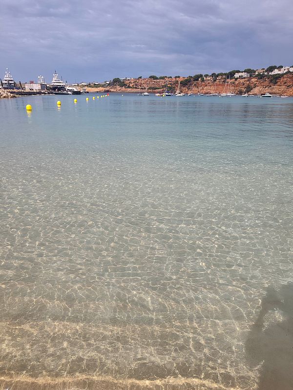 Clear shallow water at a beach with yellow buoys marking a path, boats and yachts anchored further out, and a cliffside with buildings in the background under a cloudy sky.