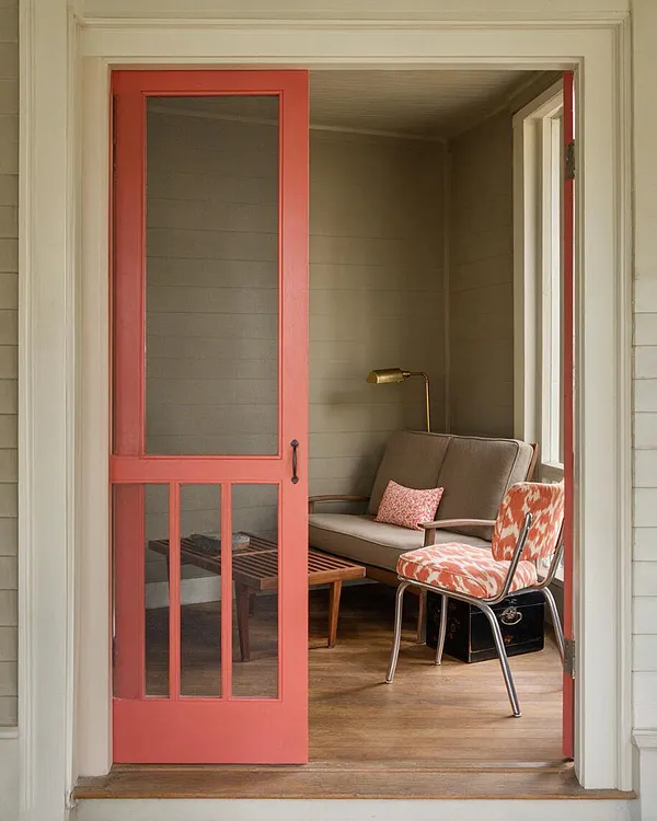 A front porch seating nook viewed through a coral-painted screen door, featuring a taupe loveseat, a patterned orange-and-white chair, a slatted wood coffee table, and a brass reading lamp. The porch has beige walls, a wood floor, and layered vintage-inspired accents.