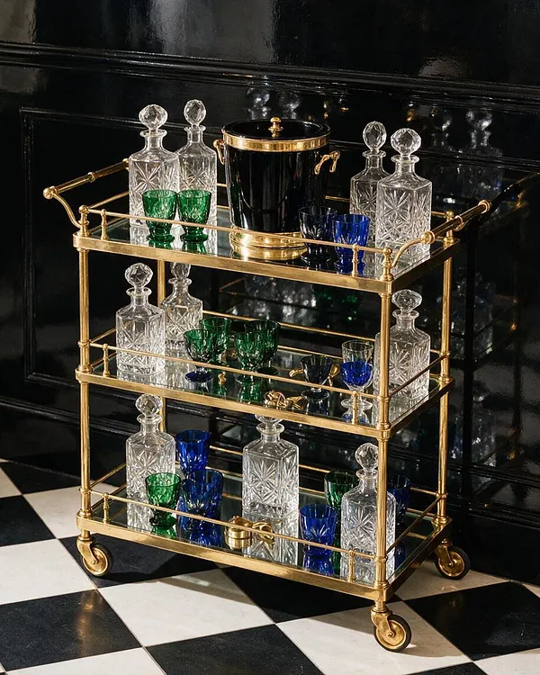 A Hollywood Regency bar cart with a polished brass frame, mirrored shelves, and small caster wheels sits against a glossy black wall. It is styled with cut crystal decanters, colorful green and blue glasses, and a black ice bucket with gold trim, creating a luxurious and dramatic display.