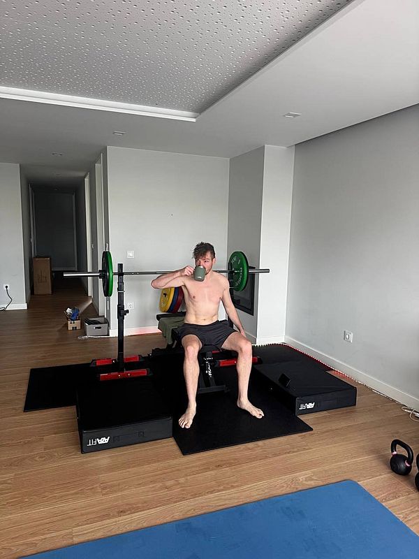 A shirtless man sits on a weight bench in a home gym, holding a drink.
