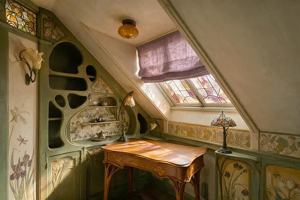 A compact attic home office with a curved Art Nouveau built-in cabinet, a carved wooden desk, and stained-glass windows under a sloped ceiling. Soft sunlight filters through a lavender Roman shade, warming the sage green woodwork, floral wall borders, and decorative glass lamp.